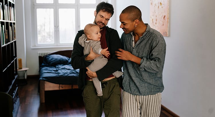 Two gay parents with a baby standing in a suburban house, looking thoughtful.